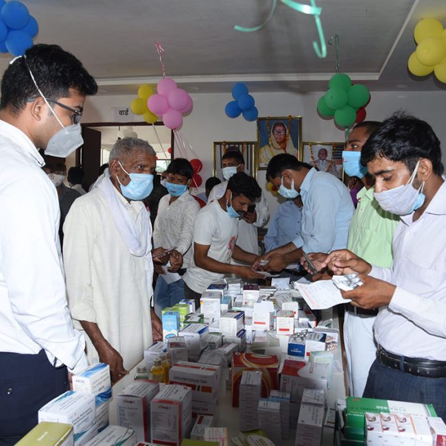 Group of medical professionals in face masks examining and distributing medicines during healthcare outreach at Ayurvedic college in UP, with colorful balloon decorations overhead"
